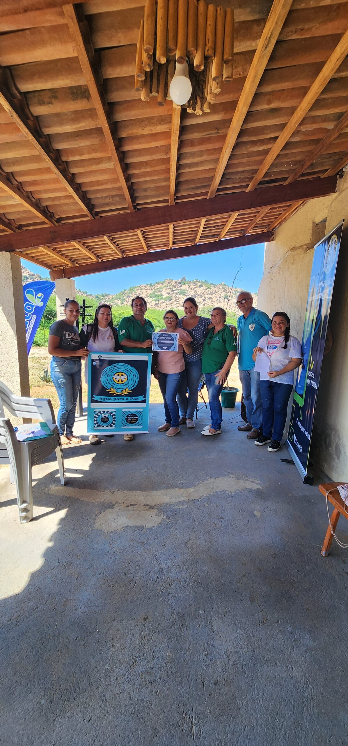 Equipe da Croatá com  diretora e professores da escola municipal Antônio Junino de Santana dos Garrotes PB. Estão no alpendre  da casa em frente à serra com banner da campanha da ONU sobre o dia mundial da água e certificado de participação.